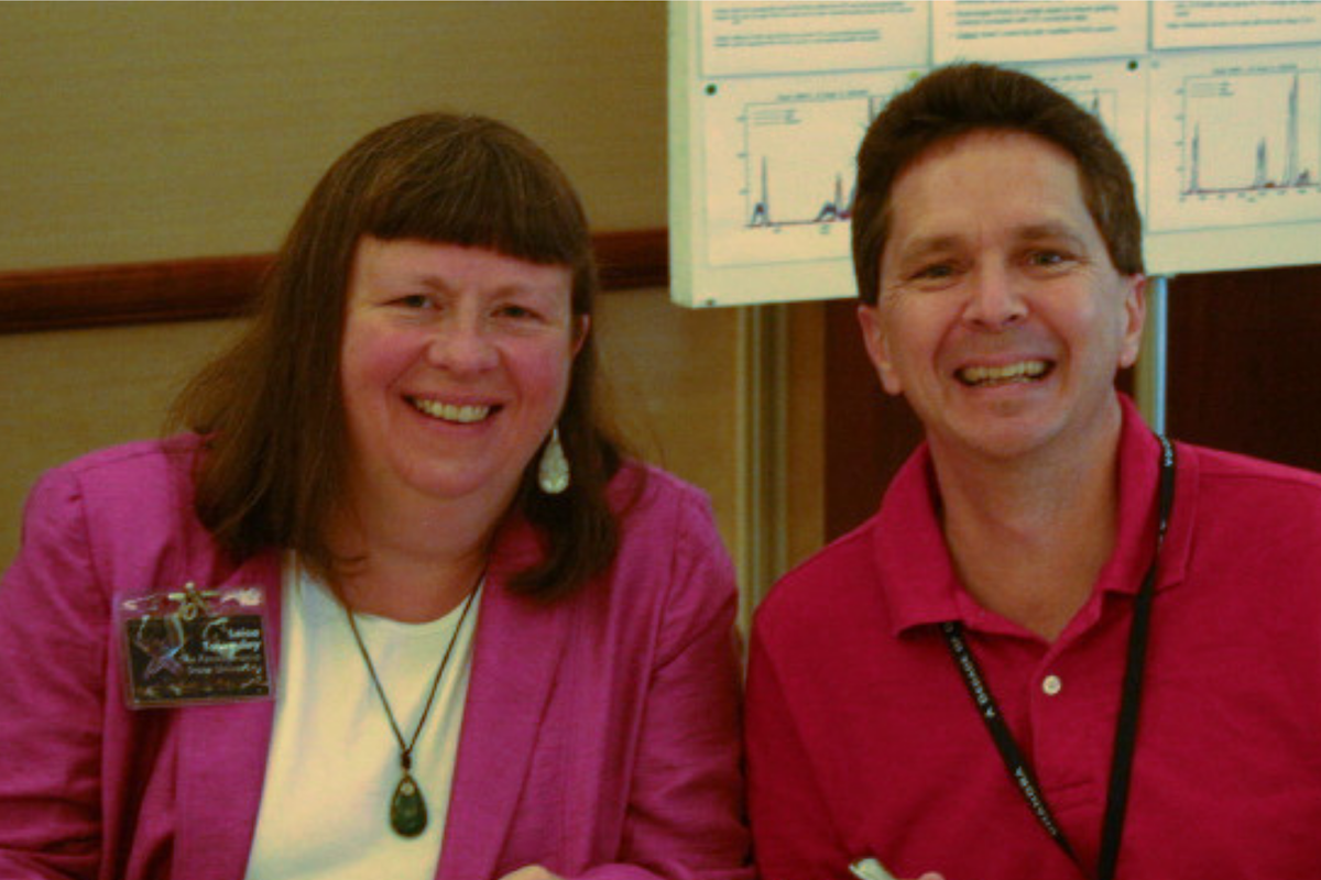 Leisa Townsley and Pat Broos smiling at the camera. Leisa has on a purple blazer and a nametag, while Pat wears a red polo with a lanyard around his neck. 