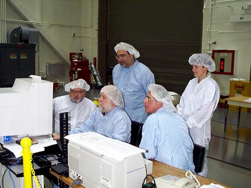 Five people in cleanroom 'bunny suits,' all focused on a CRT computer monitor. Jerry is in the rear, sitting, with his face, glasses, and beard distinguishing him from the other four people.