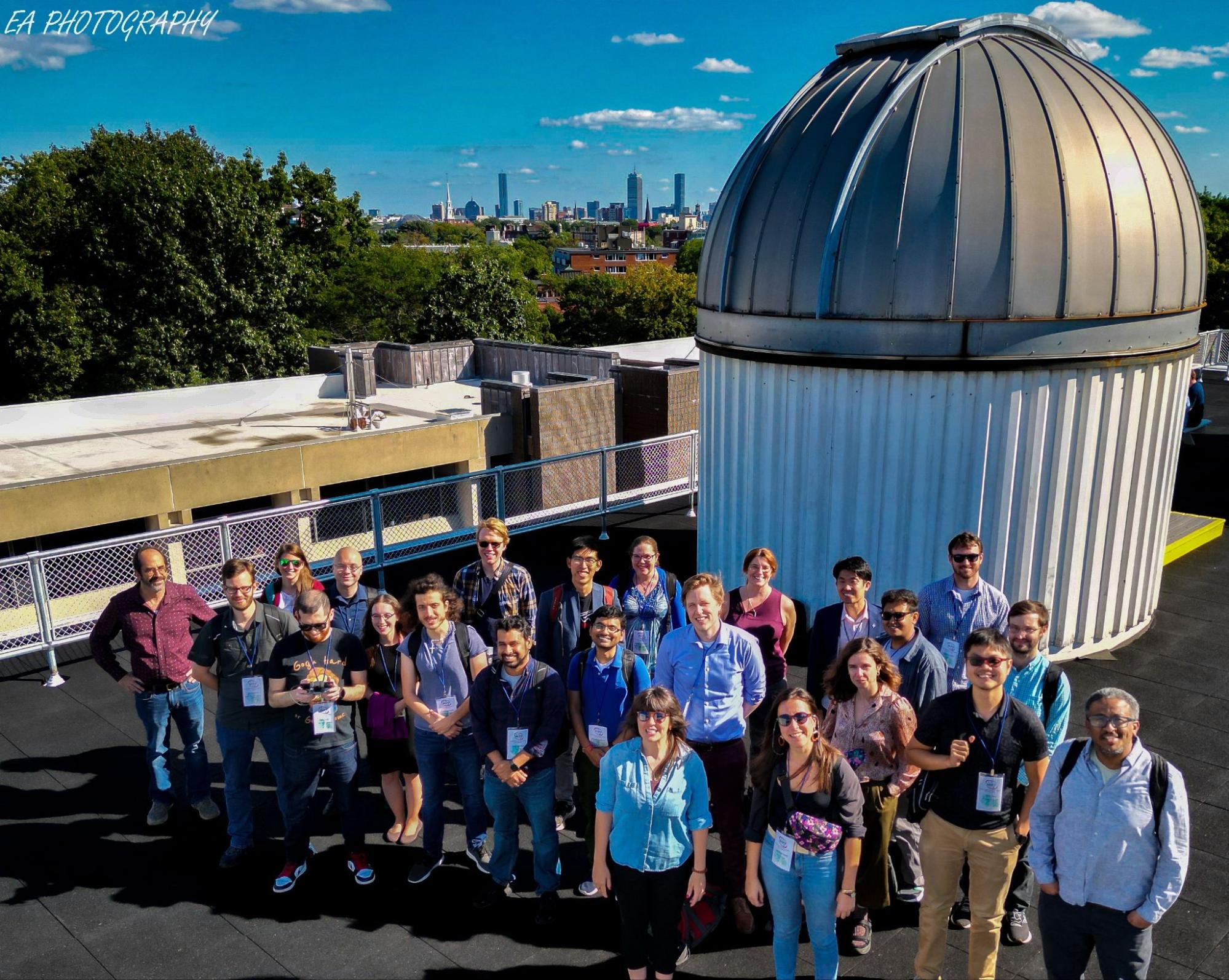 23 people standing on a roof looking upwards toward the camera. Most are dressed in business casual attire, with many carrying book bags and wearing name tag lanyards and sunglasses. Immediately behind them is a small telescope dome, several meters tall and a couple of meters wide. In the background are clear blue skies, lush green treetops, and the distant skyline of Boston.